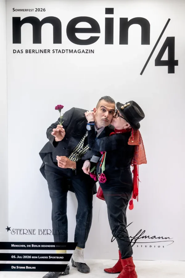 Fotobox-Aufnahme: Frau mit Zylinder küsst einen Mann im Anzug auf die Wange vor der mein/4-Wand